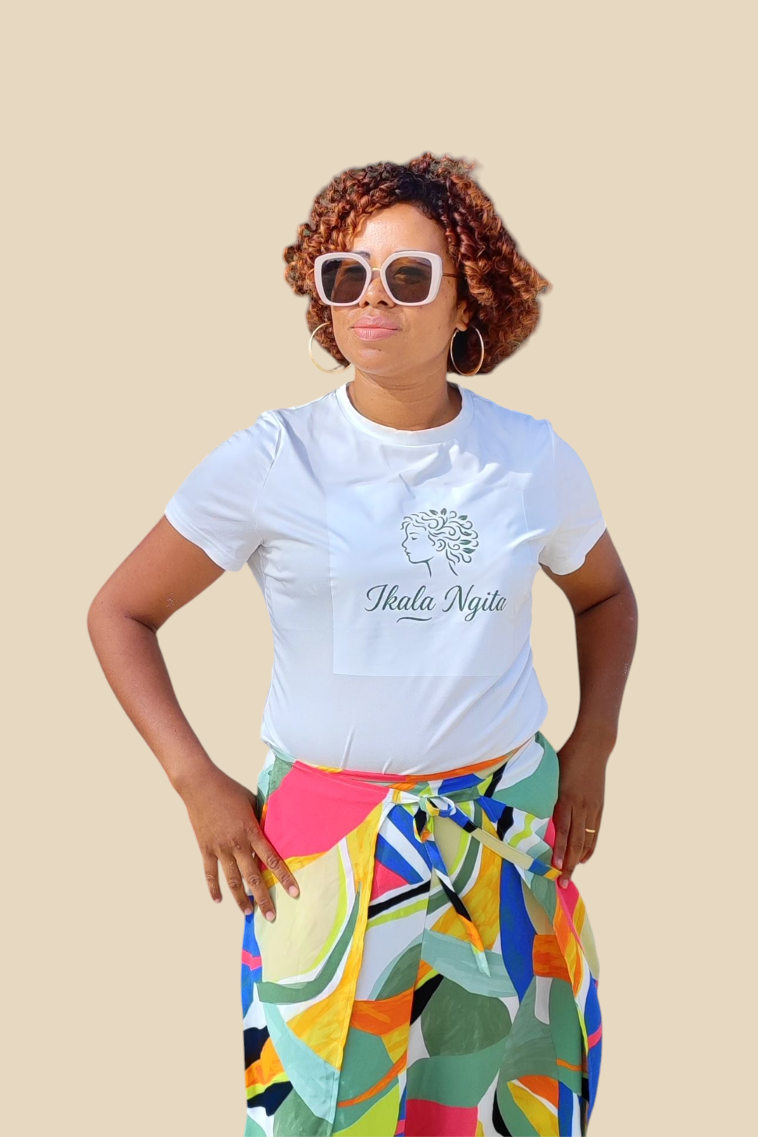 Femme portant un t-shirt blanc Ikala Ngita et un pagne coloré, sur fond beige sable. Mode africaine moderne et colorée.