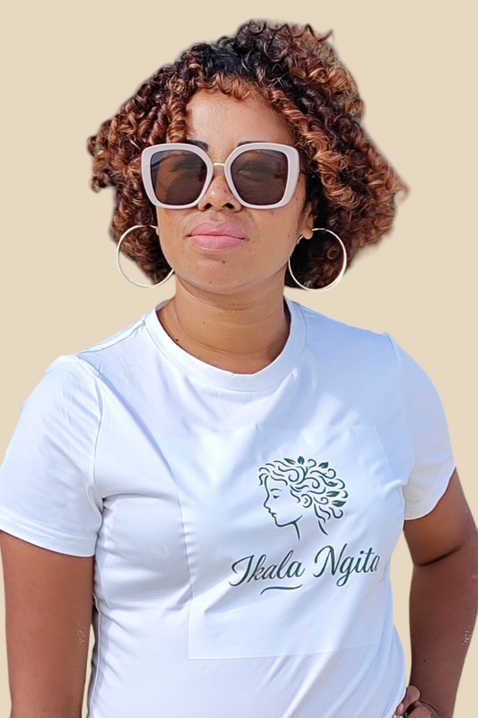Femme aux cheveux bouclés cuivrés portant un t-shirt blanc avec le logo Ikala Ngita, marque de soins capillaires naturels pour cheveux texturés. Look estival avec grandes lunettes de soleil, incarnant l’élégance et l’identité afro-malgache