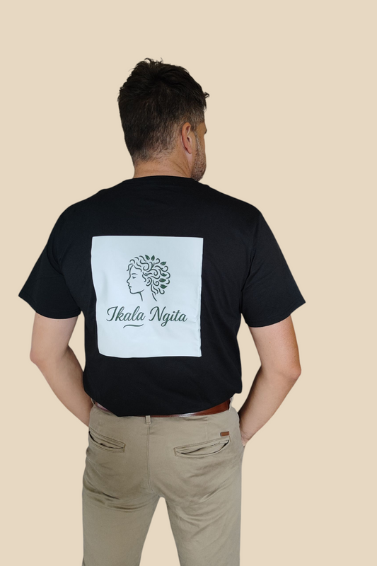 Homme vu de dos portant un t-shirt noir Ikala Ngita avec logo blanc et un pantalon beige, sur fond beige sable. Mode élégante et décontractée.