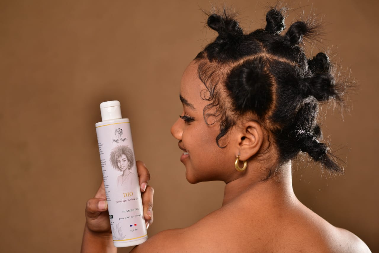 Jeune femme aux cheveux texturés coiffés en bantu knots tenant un shampoing Dio d’Ikala Ngita, mettant en avant une routine adaptée aux cheveux crépus et texturés. 