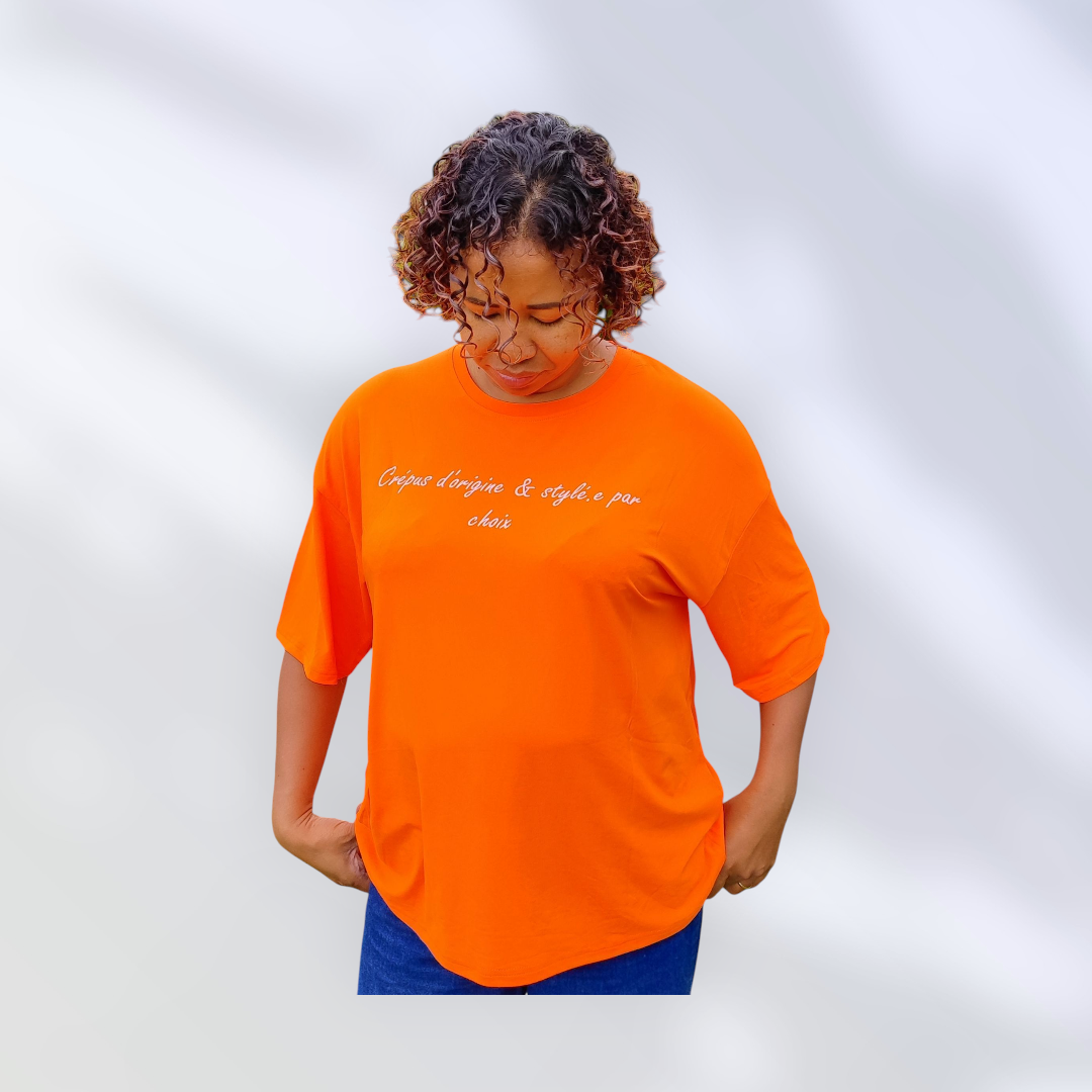 personne de face portant un tee-shirt orange avec l'inscription « Crépus d’origine & stylée par choix .